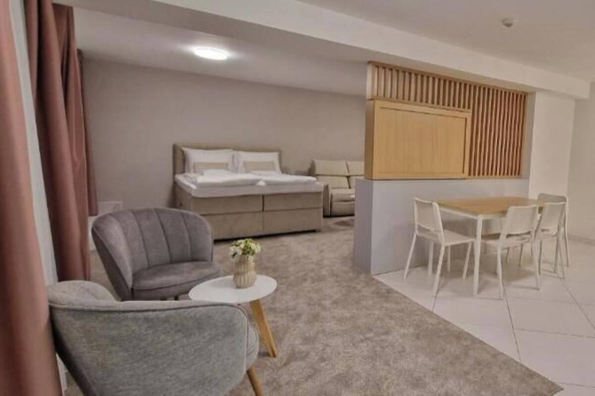 Ein modernes Hotelzimmer mit Bett, einem kleinen Esstisch mit Stühlen, einem grauen Sessel, einem runden Beistelltisch und einer Küchenzeile.