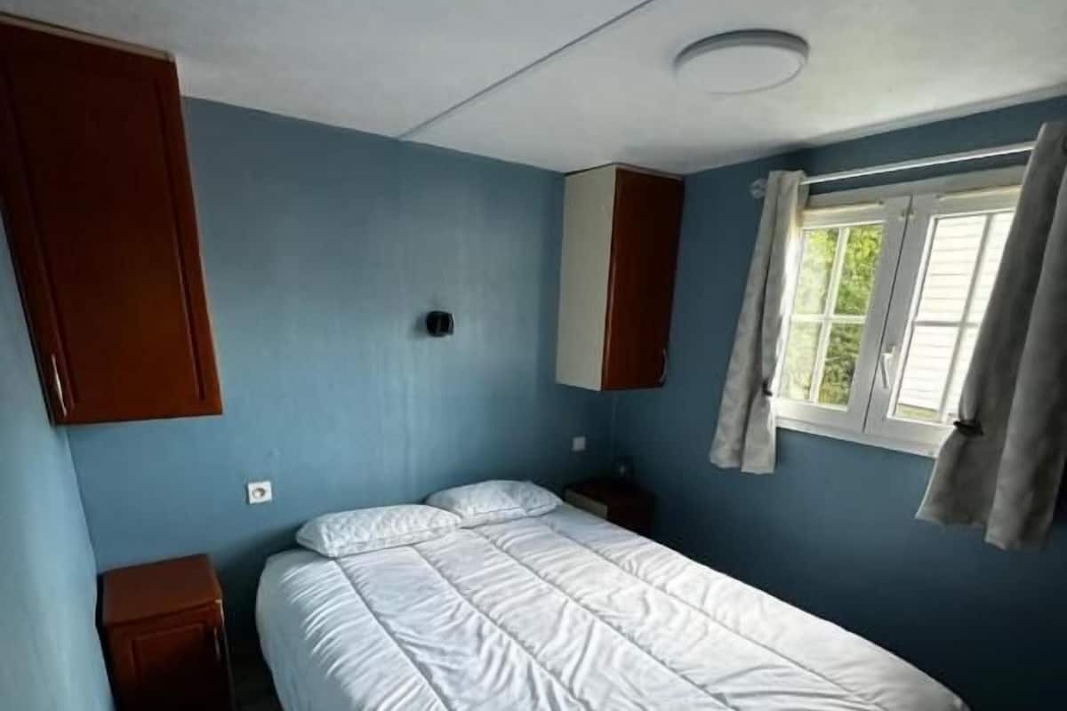 A single-bed room with a wooden wardrobe, a small bedside table, and a window with curtains.