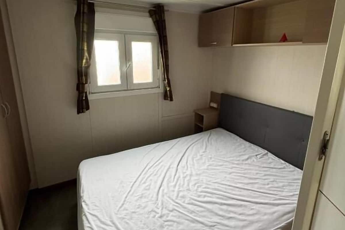 A single-bed room with a window, curtains, a bedside table, a shelf, and a wardrobe.