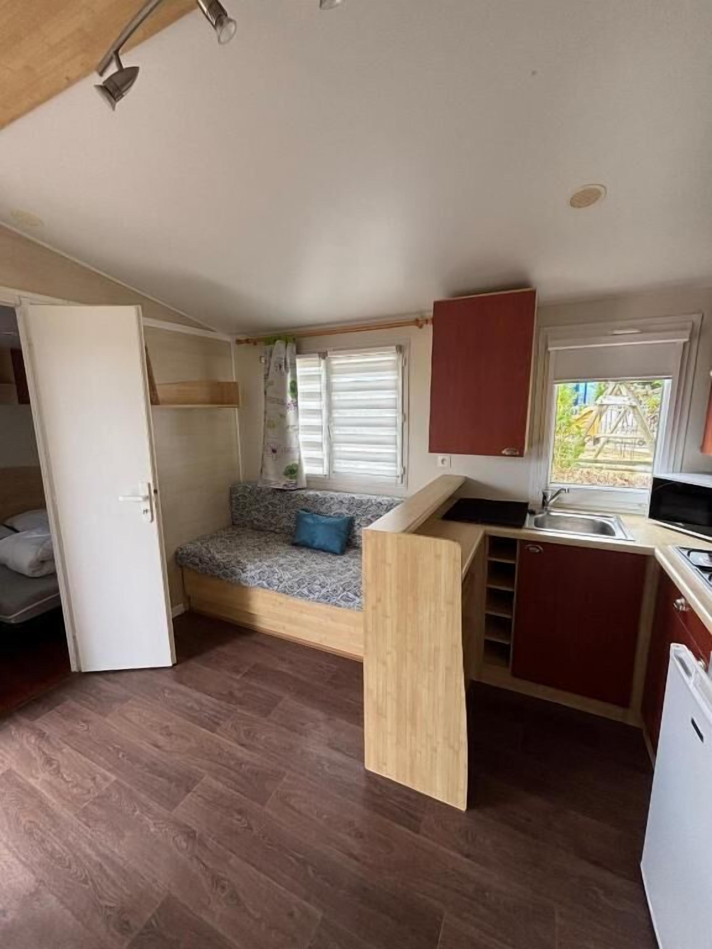 A compact RV interior with a kitchenette, a sleeping area with a bed, and a window with blinds.