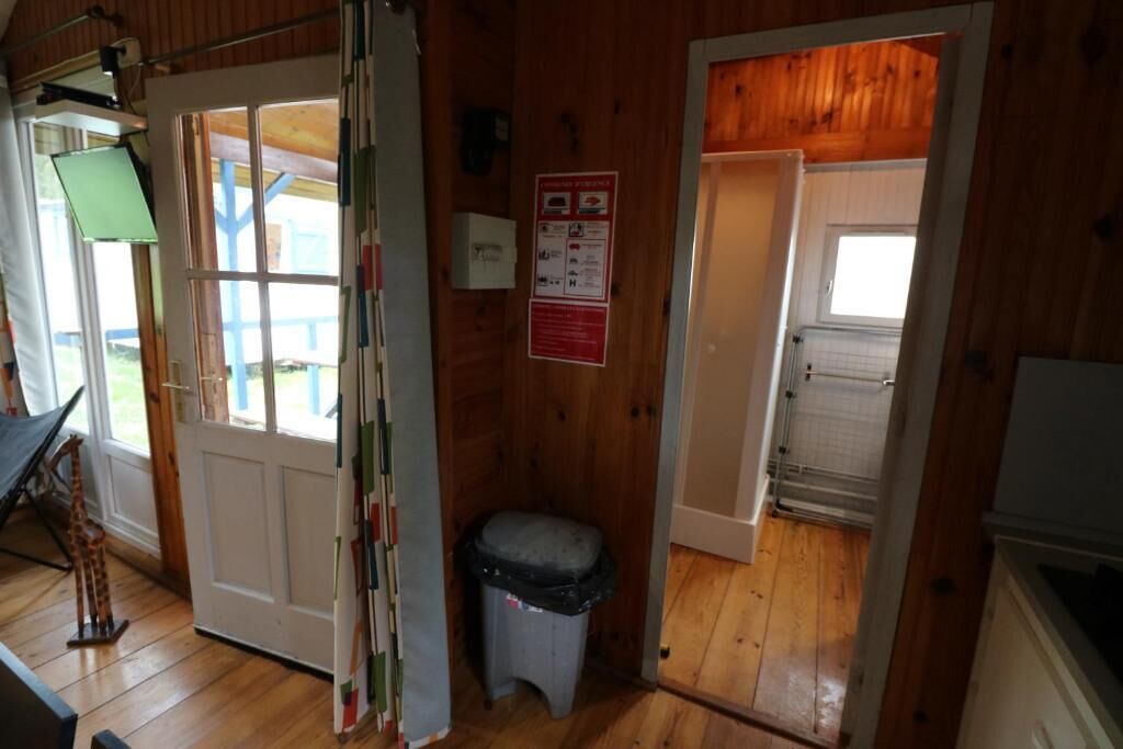 Una cabaña de madera con una puerta que da a una habitación pequeña, una ventana y un contenedor de basura.