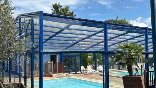 Uma área de piscina coberta com estrutura de vidro, cadeiras de espreguiçadeira e uma mesa pequena.