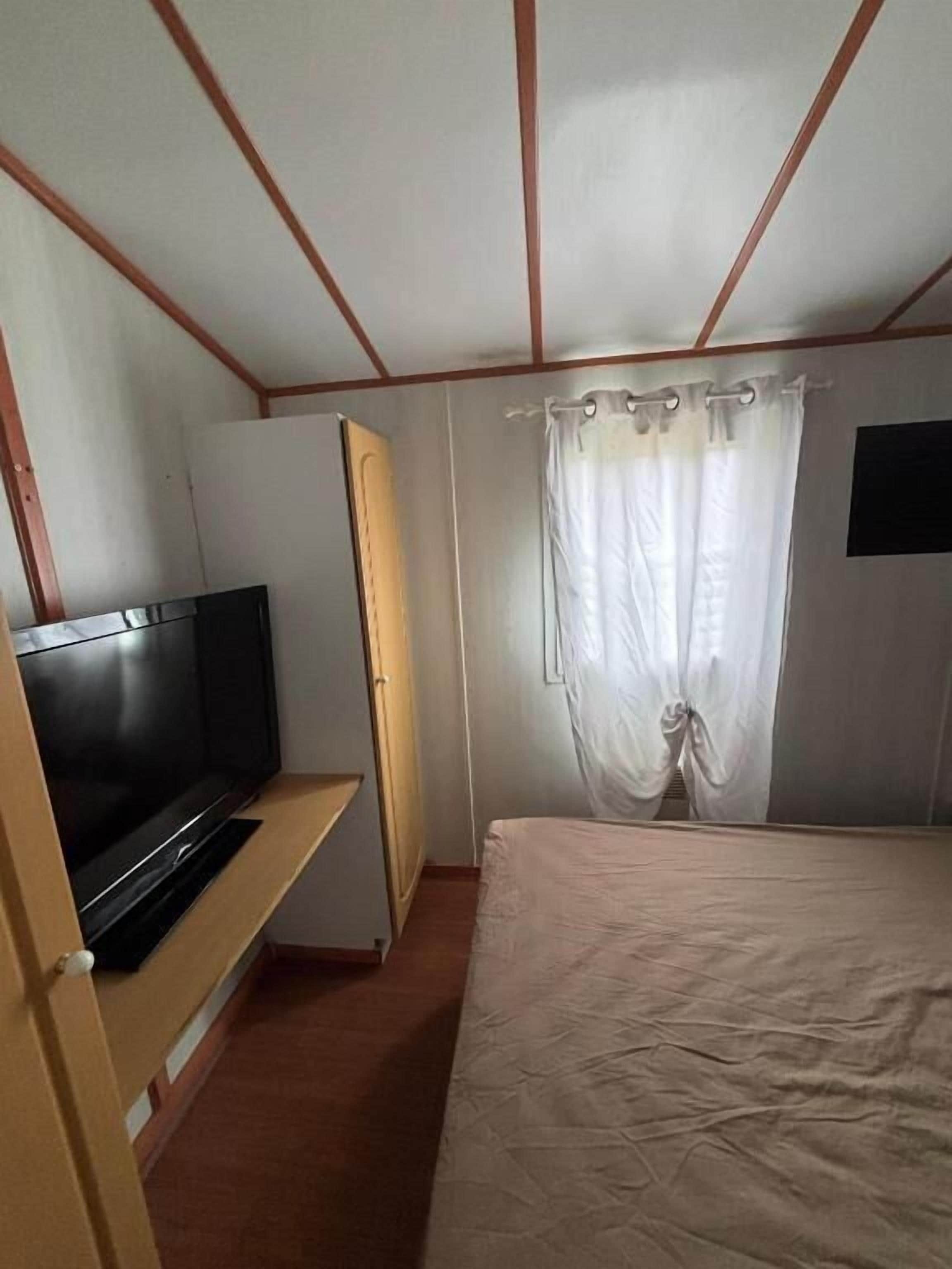 Un dormitorio con cama, televisor sobre un mueble, armario y ventana con cortinas.