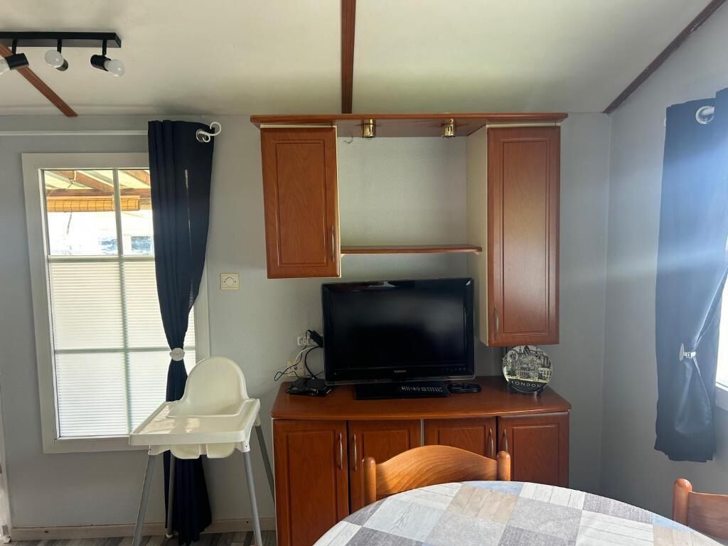 Une petite pièce avec un téléviseur posé sur un meuble en bois, une chaise blanche et une table recouverte d’une nappe à carreaux.