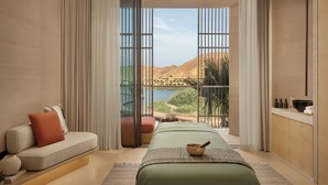 Een kamer met een bed, een massagetafel, een bank en uitzicht op een wateroppervlak, plus een balkon met een leuning.