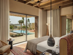 A bedroom with a large window, a bed, a bedside table, a lamp, and a view of an outdoor swimming pool.