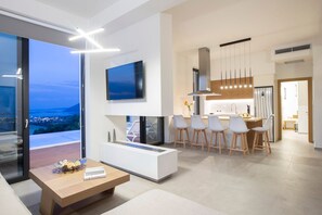 A modern living room with a large window, a flat-screen TV, a dining area with bar stools, and a kitchen with a stainless steel refrigerator. - Villa Karyatis, Divine Sea & City Views, Infinity Pool, Furnished Terrace,. (Lefkada)
