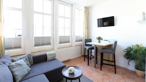 A modern living room with a sofa, a dining table with two chairs, a TV mounted on the wall, and a potted plant. - Apartment no. 01 (Warnemünde)