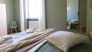 Una camera da letto con un letto, una sedia, un termosifone, una finestra con tende e uno specchio a parete.