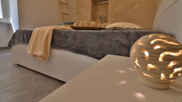 A modern bedroom with a stone wall, a bed with a grey headboard, a wooden ladder, and a decorative light fixture. - Vacation Home in Otranto with 2 bedrooms sleeps 4 (Otranto)