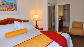 Habitación de hotel con una cama grande, una mesita de noche con lámpara, una silla y un baño visible a través de una puerta abierta.