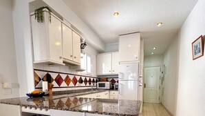 Una cocina moderna con armarios blancos, encimera de granito y un refrigerador de vinos empotrado. Hay tres taburetes de bar de madera con asientos acolchados.