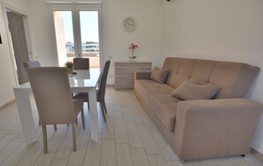 Ein modernes Wohnzimmer mit einem beigen Sofa, einem Esstisch mit Stühlen, einer kleinen Topfpflanze und einer Wanduhr.