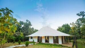 Um edifício em forma de tenda, de cor branca e com telhado plano, rodeado por um jardim com arbustos e árvores diversas.