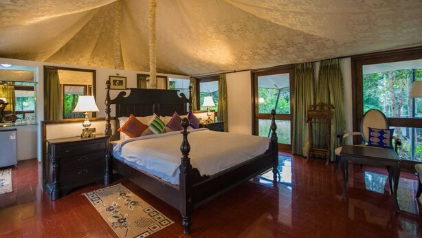 Quarto espaçoso com uma cama grande, penteadeira de madeira, escrivaninha e cadeira. Várias janelas oferecem luz natural e vista para o exterior.