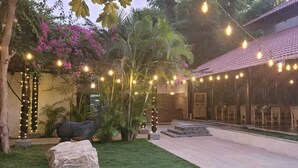 Uma área de estar ao ar livre coberta, decorada com luzes de corda, uma escultura de pedra e muita vegetação.