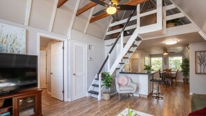 A spacious living area with a wooden floor, a large flat-screen TV, a staircase with a white railing, and a dining area with a table and chairs. - Lake Henry A-Frame Retreat-Dock, Kayaks & Fire Pit (Lake Placid)