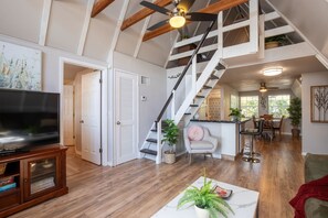 Un salón amplio con suelo de madera, un televisor de pantalla plana grande, una escalera con barandilla blanca y un comedor con mesa y sillas.