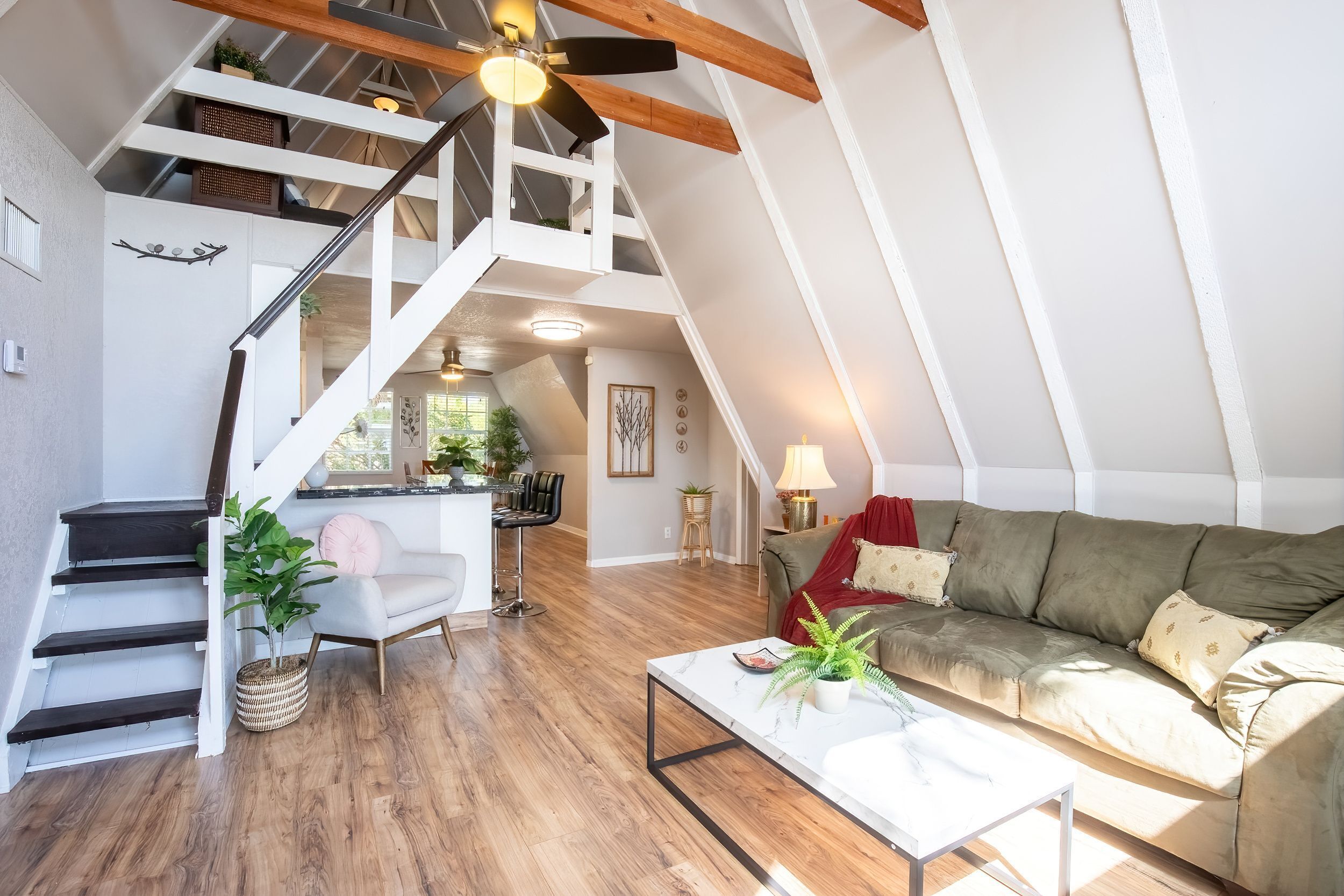Un loft amplio con techo alto, vigas de madera, una escalera, un sofá verde, una mesa de centro blanca y un comedor.
