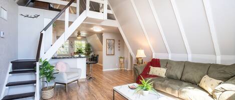 Un loft amplio con techo alto, vigas de madera, una escalera, un sofá verde, una mesa de centro blanca y un comedor.