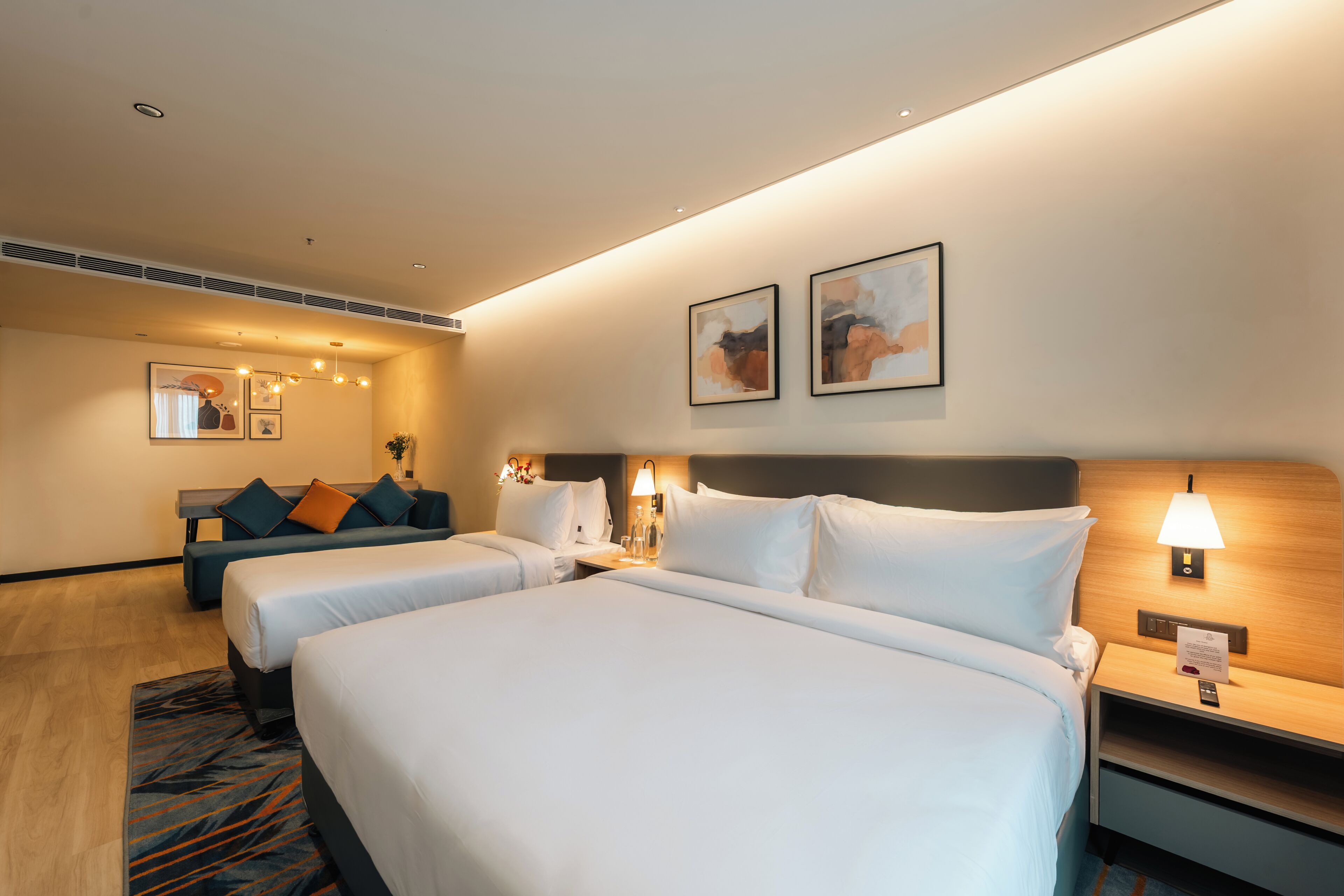 A hotel room with two beds, a sofa, a nightstand with a lamp, and artwork on the walls.