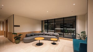 Un hall d’entrée moderne avec un canapé d’angle, deux tables rondes et une grande fenêtre donnant sur un jardin intérieur.