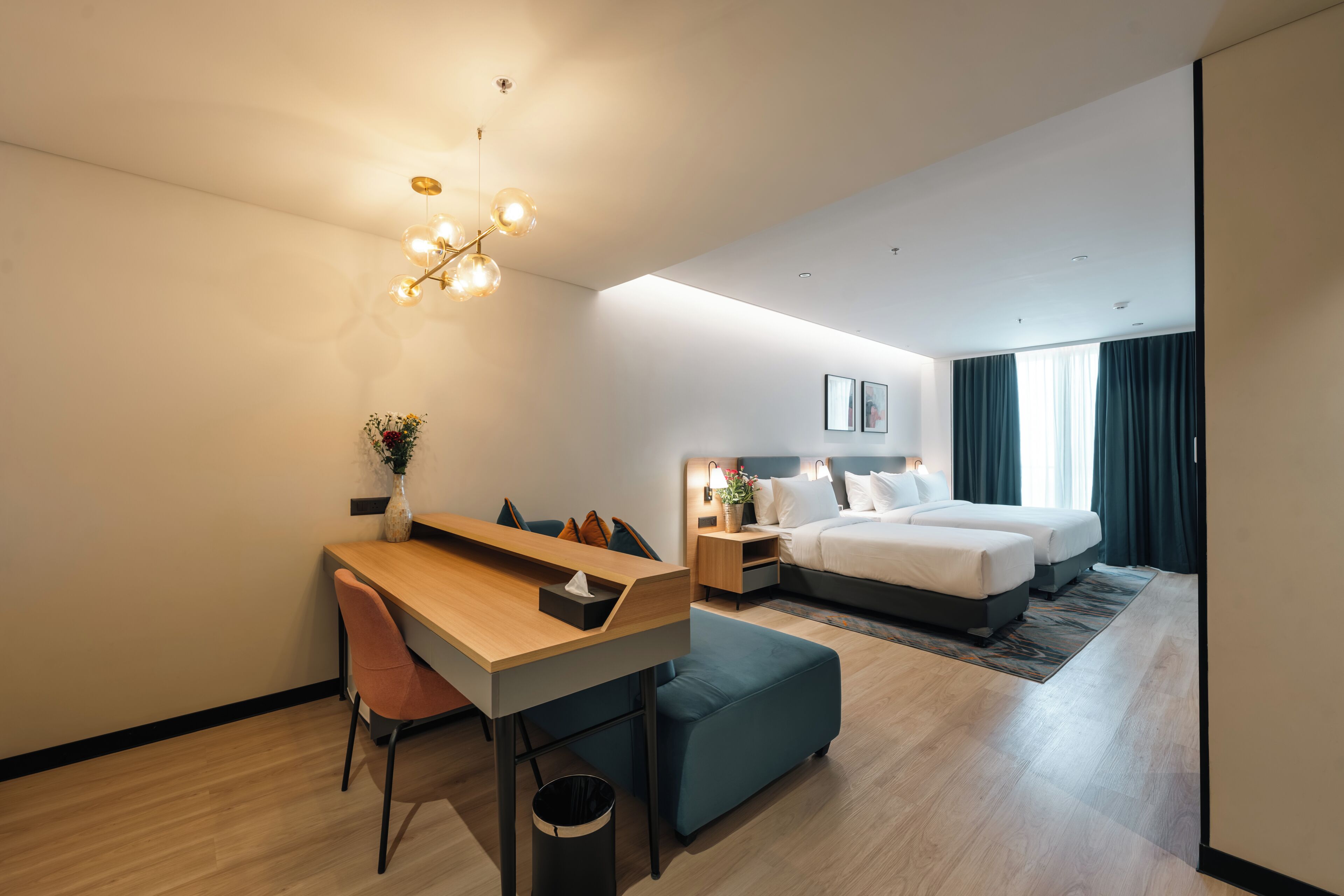 Ein modernes Hotelzimmer mit Schreibtisch aus Holz, blauem Hocker, einem Bett mit zwei Kissen, einem Nachttisch mit Lampe und einem Wandbild.