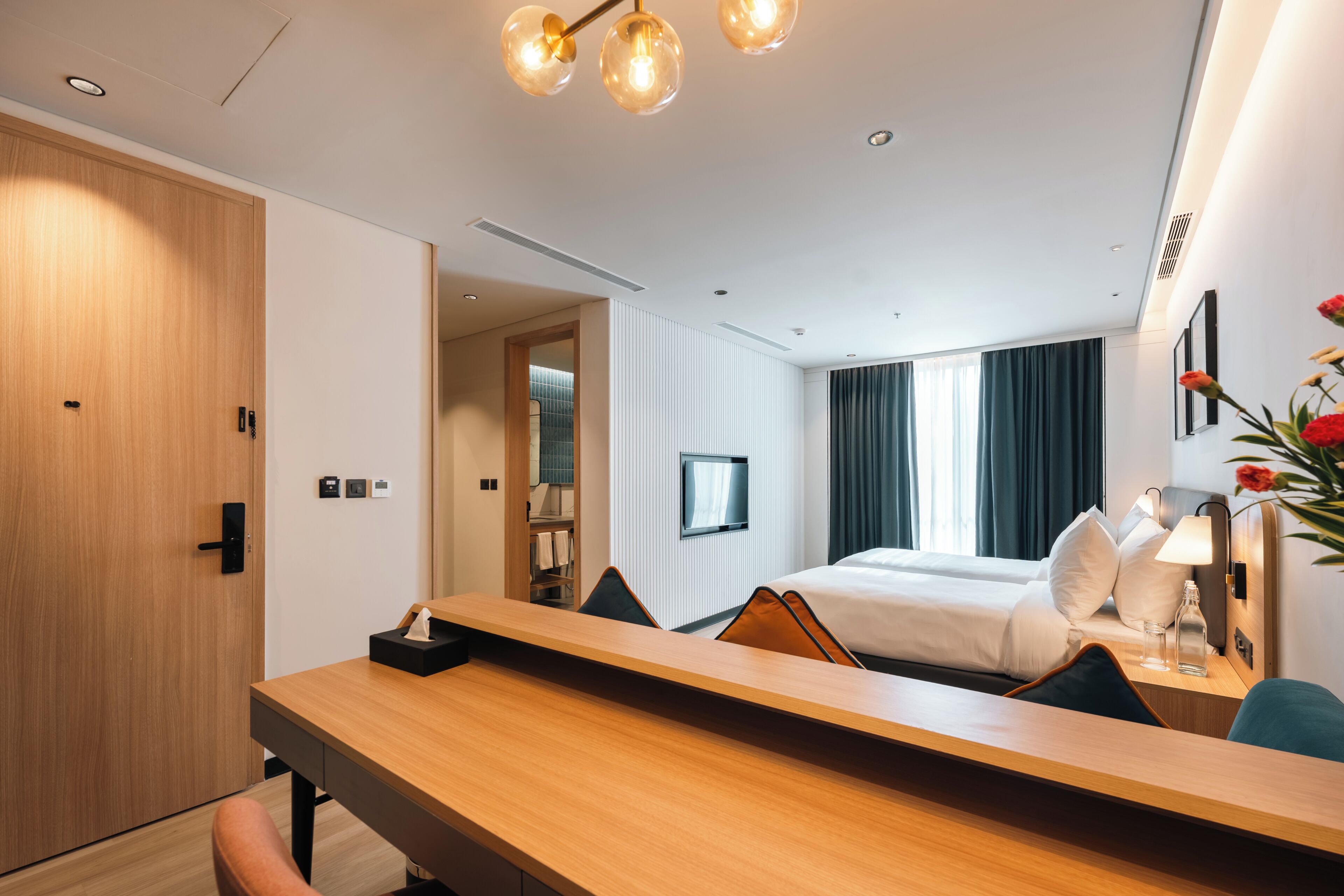 A hotel room with a wooden desk, a bed with white linens, a TV on the wall, and a large window with curtains.
