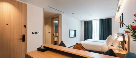 A hotel room with a wooden desk, a bed with white linens, a TV on the wall, and a large window with curtains.