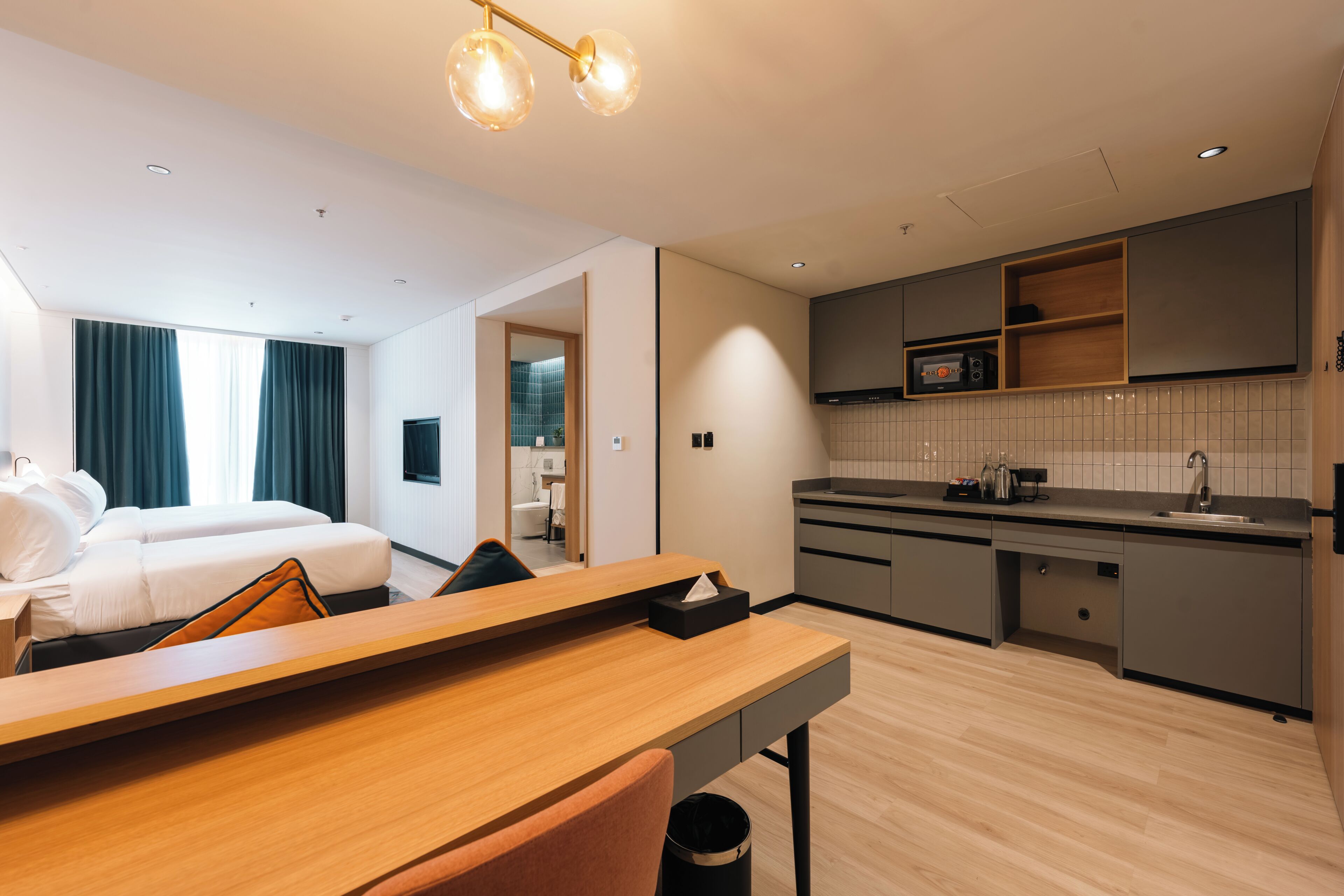 A hotel room with a wooden desk, two beds, a kitchenette, and a bathroom visible through an open door.