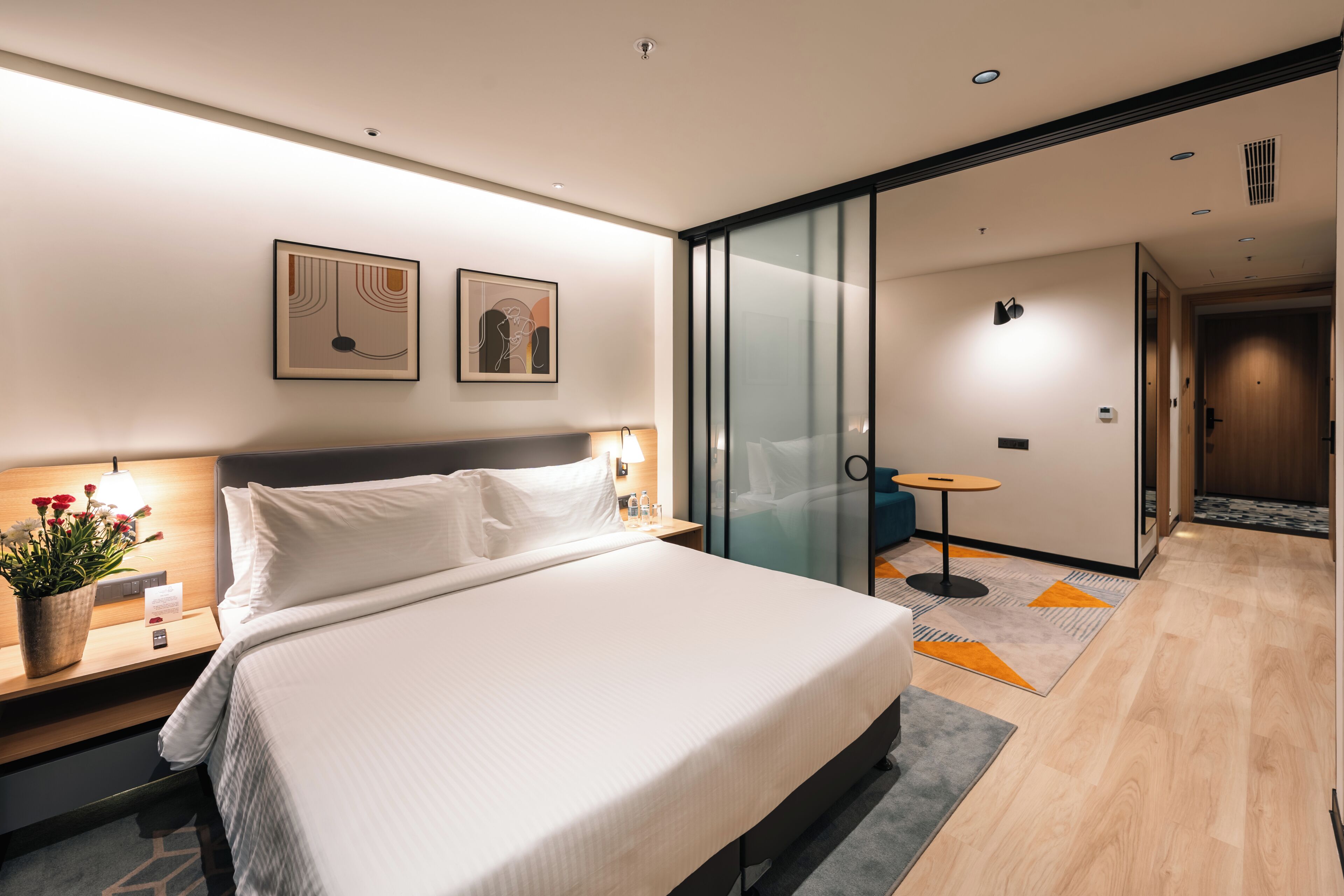 Una habitación de hotel moderna con una cama grande, mesitas de noche, una mesita auxiliar con una lámpara, una planta en maceta y obras de arte en las paredes.