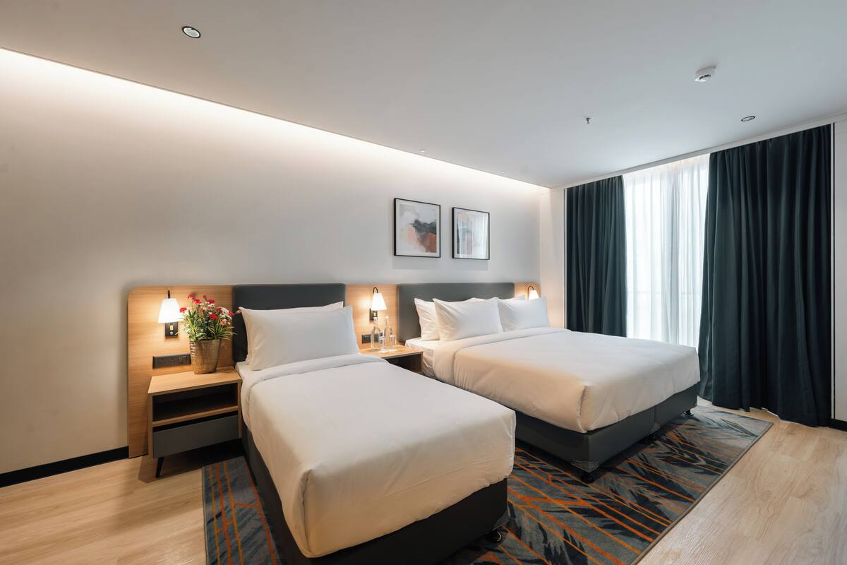Hotellihuone, jossa on kaksi sänkyä, yöpöytä lampun kanssa, pieni pöytä maljakolla ja ikkuna, jossa on verhot.