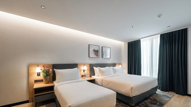 Une chambre d’hôtel avec deux lits, une table de chevet avec une lampe, une petite table avec un vase et une fenêtre avec des rideaux.