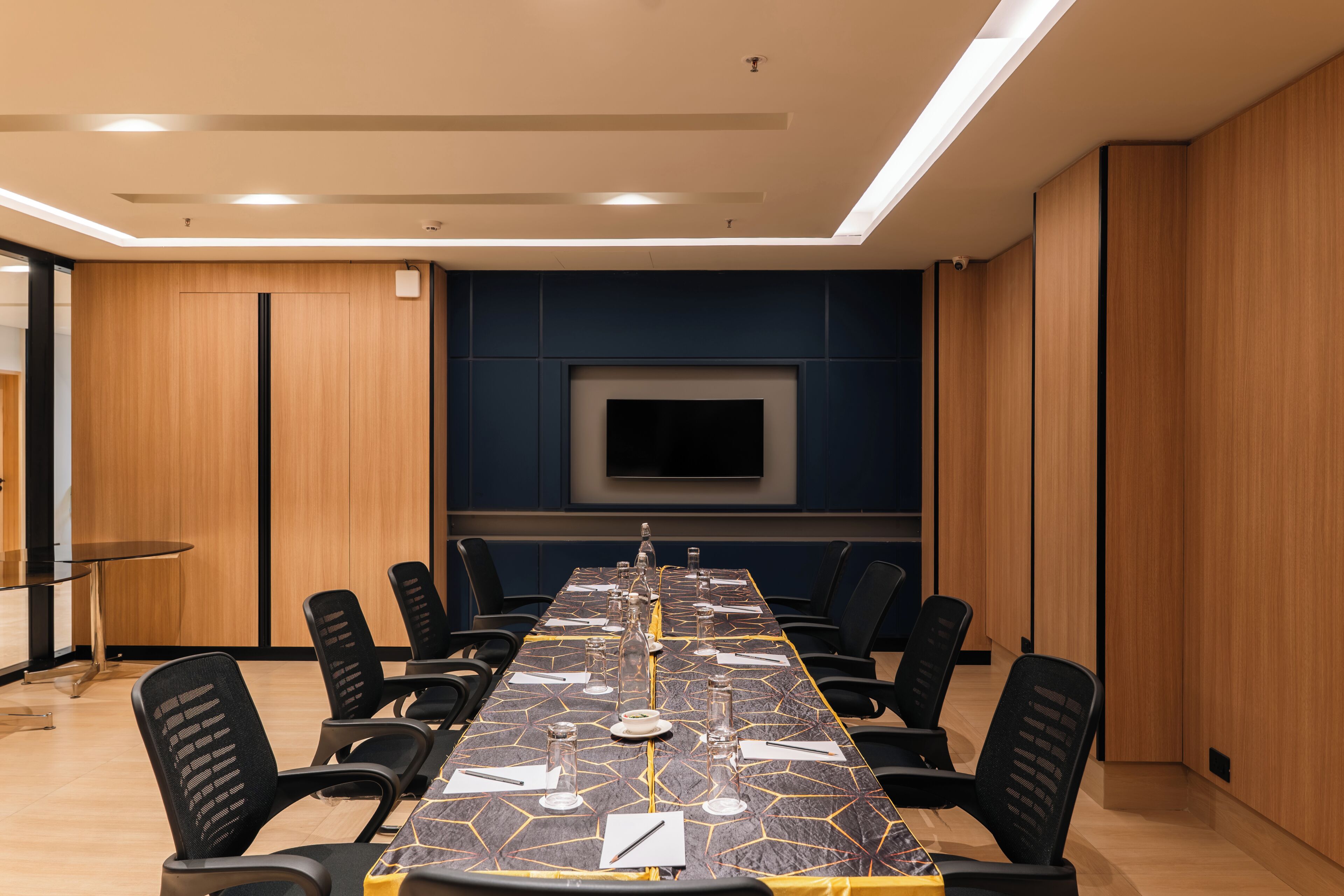 A conference room with a long table, chairs, a flat-screen TV, and wooden paneling.