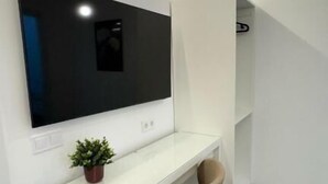 A modern, white room with a large flat-screen TV mounted on the wall, a white desk, a beige chair, and a small potted plant. - jacuzzi rooms (Madrid)