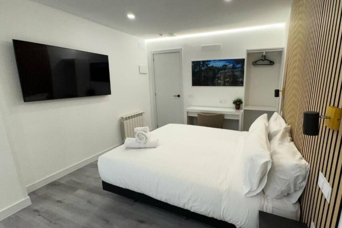 A modern hotel room with a large bed, a flat-screen TV mounted on the wall, a desk with a chair, and a mirror.