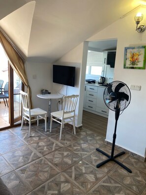 A dining area with a table and chairs, a TV mounted on the wall, a fan, and a kitchen in the background.