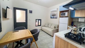 A compact living space with a kitchenette, a dining area with a wooden table and chairs, a sofa, and a small TV. - Paddys Pansiyon (Fethiye)