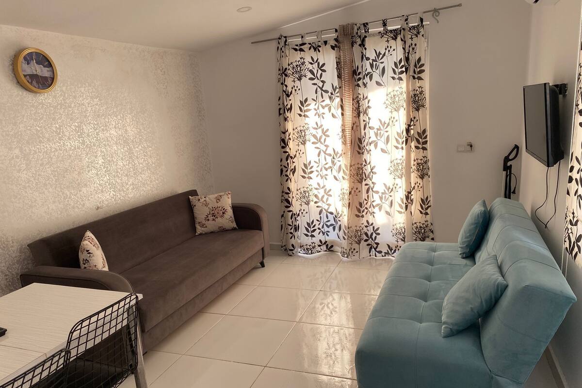 A living room with a brown sofa, a blue sofa, a dining table with chairs, and a TV mounted on the wall.