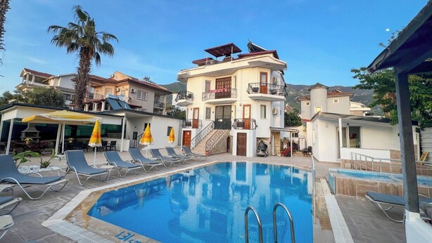 A swimming pool with lounge chairs and umbrellas, surrounded by buildings with balconies and a mountain in the background. - Paddys Pansiyon (Fethiye)