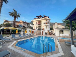 A swimming pool with lounge chairs and umbrellas, surrounded by buildings with balconies and a mountain in the background. - Paddys Pansiyon (Fethiye)