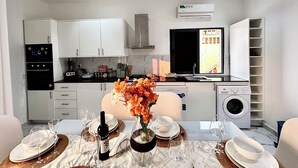 Een kleine keuken met witte kastjes, een inbouwoven en een magnetron. Er is een eethoek met een tafel gedekt voor twee personen, een vaas met bloemen en een fles wijn.
