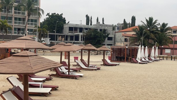 Een strand met rijen ligbedden en houten parasols, omringd door gebouwen en palmbomen.