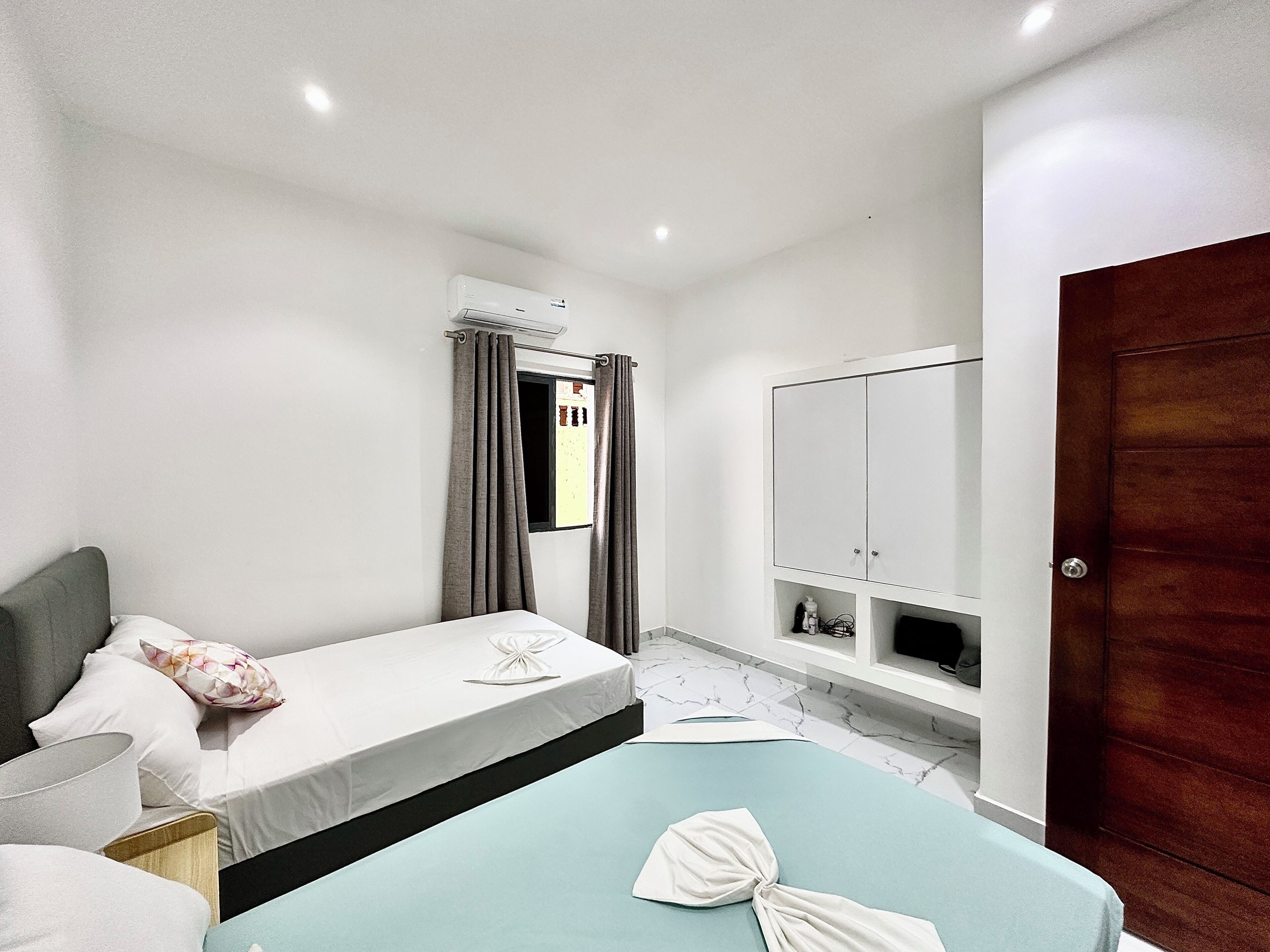 A bedroom with two beds, a wardrobe, a window with curtains, and an air conditioning unit.