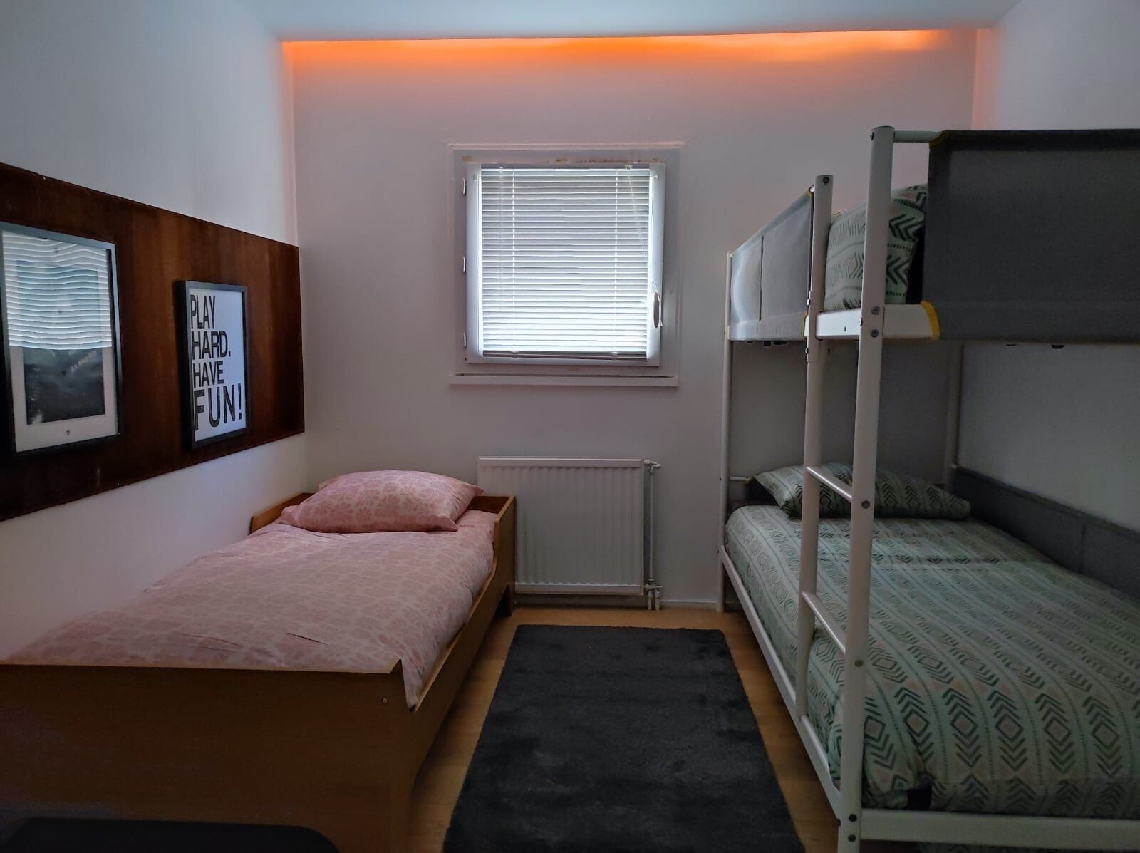 Un dormitorio pequeño y bien iluminado con una cama individual, una litera, un radiador, una ventana con persianas y dos cuadros en la pared.