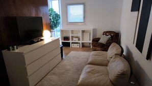 Olohuoneessa on beigenvärinen sohva, valkoisessa kaapissa oleva televisio ja ikkuna, jossa on sälekaihtimet.