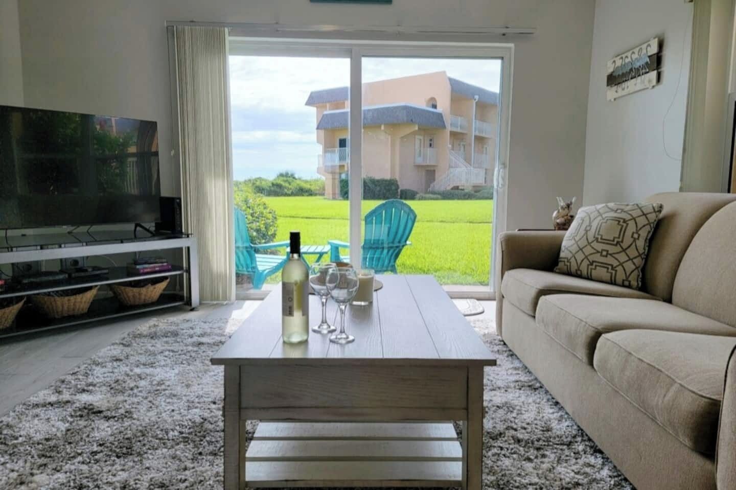 Un salon avec un téléviseur, une table basse en bois sur laquelle se trouvent une bouteille et des verres, un canapé et une vue sur un immeuble à travers de grandes fenêtres.