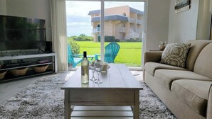 Un salon avec un téléviseur, une table basse en bois sur laquelle se trouvent une bouteille et des verres, un canapé et une vue sur un immeuble à travers de grandes fenêtres.