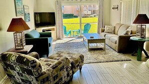 Un salon comprenant un canapé, un fauteuil et une table basse, une télévision, et donnant sur un champ jaune par une porte vitrée.