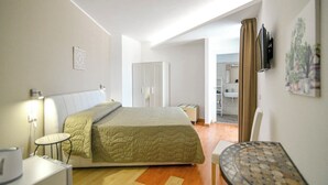 Et hotelværelse med seng, lille bord, stol, skrivebord, fjernsyn og et badeværelse, der ses gennem en åben dør.